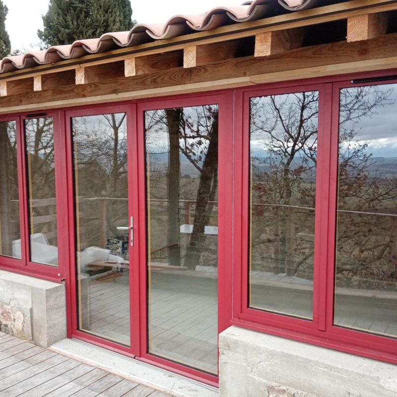 Baies vitrées rouges modernes ouvrant sur une terrasse en bois, offrant une vue panoramique sur un paysage boisé sous un toit en tuiles.