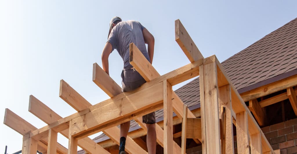 Ouvrier en casquette sur une charpente en bois, construisant un ajout de maison sous ciel clair. Toit en bardeaux bruns visible.