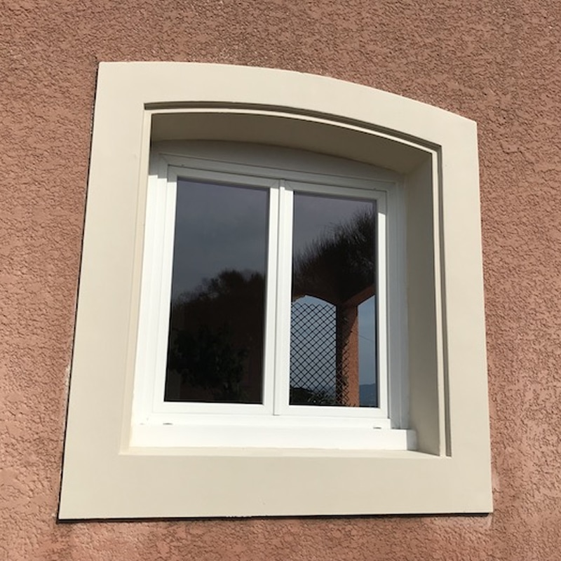 Fenêtre blanche à double vitrage sur mur texturé ocre. Encadrement beige clair avec arc discret au-dessus. Reflets du ciel et grillage.