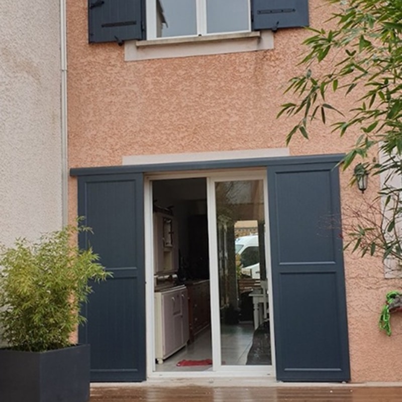 Extérieur maison mur pêche, grande porte coulissante vitrée et volets gris anthracite. Fenêtre et volets à l'étage. Plantes décoratives.