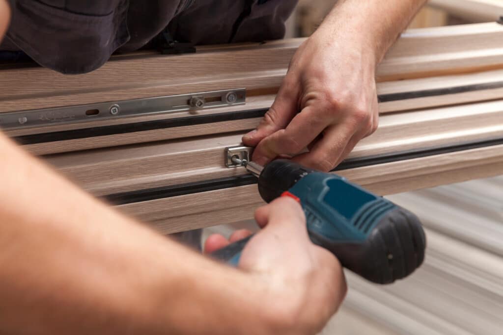 Un ouvrier installe la quincaillerie d'une fenêtre en bois clair avec une visseuse électrique, fixant une vis dans le dormant.