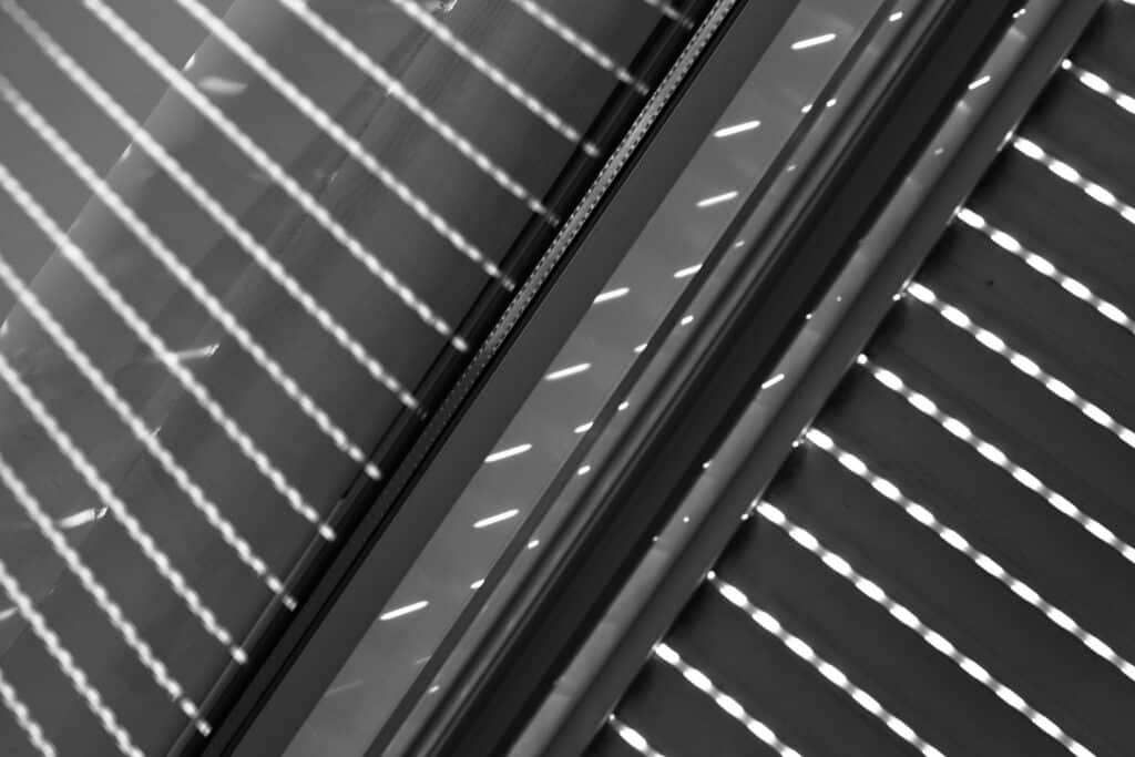 Lignes lumineuses diagonales traversant un espace sombre, projetées par des stores vénitiens en noir et blanc, créant un motif géométrique abstrait.