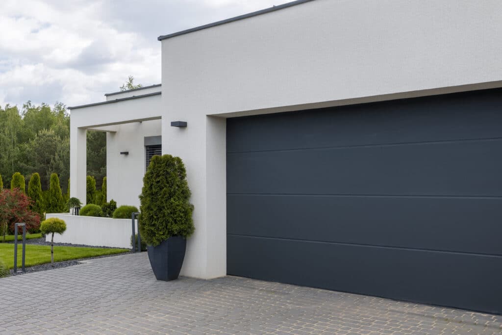 Maison moderne grise et blanche avec porte de garage anthracite. Allée pavée, jardin paysager vert et ciel nuageux.