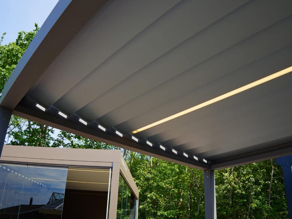 Pergola bioclimatique grise avec lattes ajustables, bande lumineuse LED jaune et spots encastrés. Arbres verts et ciel bleu en fond.