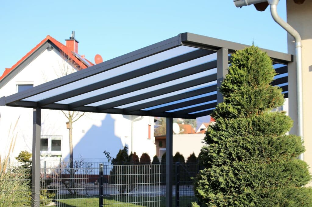 Pergola moderne grise à toit translucide devant une maison blanche aux tuiles rouges sous ciel bleu. Jardin avec grand conifère et clôture métallique.