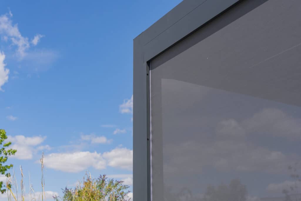 Coin de pergola moderne grise avec stores latéraux en tissu sous un ciel bleu nuageux. Végétation verte en arrière-plan.