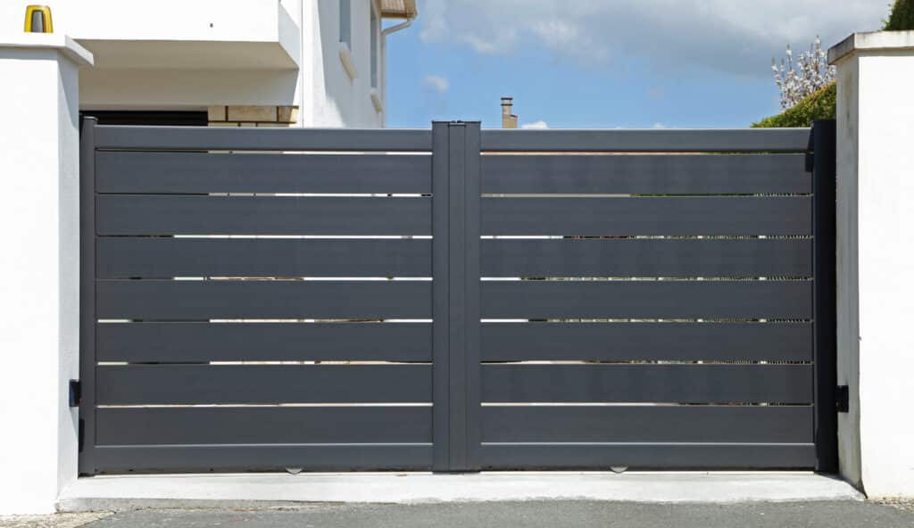 Modern gris anthracite coulissant aluminium gate avec capteurs sécurité, feu clignotant et roues visibles. Maison et ciel bleu en arrière-plan.