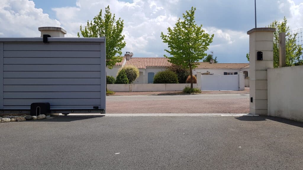 Portail coulissant gris ouvert sur rue résidentielle. Maisons aux toits tuilés, arbres verts et ciel nuageux. Le numéro 3 est visible.