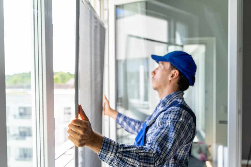 Technicien en casquette bleue installant une moustiquaire ou un store sur une fenêtre lumineuse, vue urbaine floue.