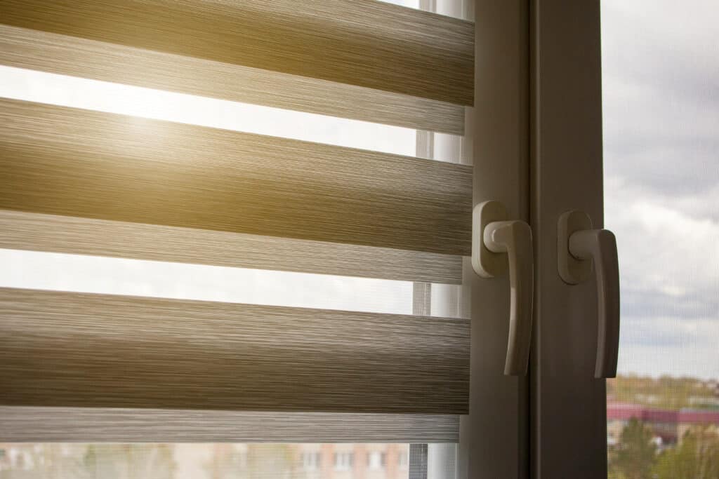 Store jour-nuit beige rayé occultant la lumière du soleil à travers une fenêtre. On voit le cadre blanc et des poignées, avec l'extérieur flou.