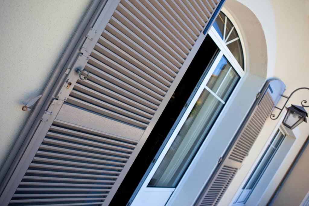 Volet gris à lattes ouvert sur une fenêtre arquée sur mur crème. Détails de quincaillerie et lanterne décorative.