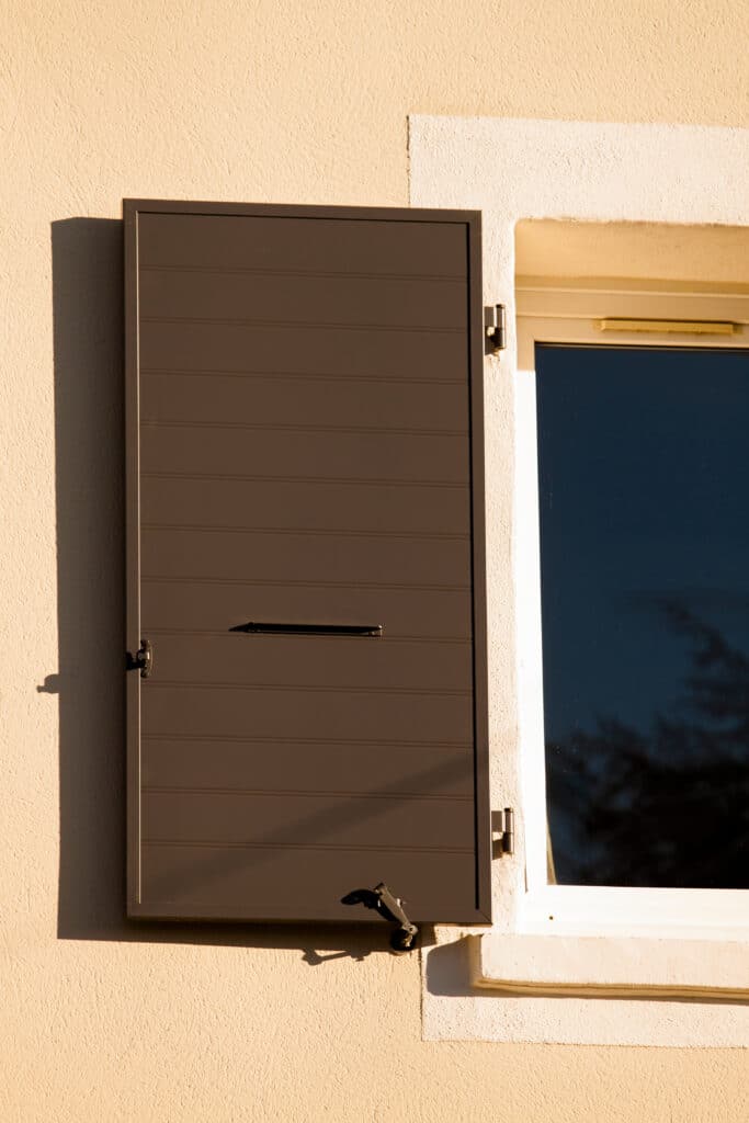 Volet marron fermé sur façade beige texturée, avec poignée et loquet noirs. Lumière du soleil intense, ombres nettes. Fenêtre blanche à côté, reflet d'arbre.