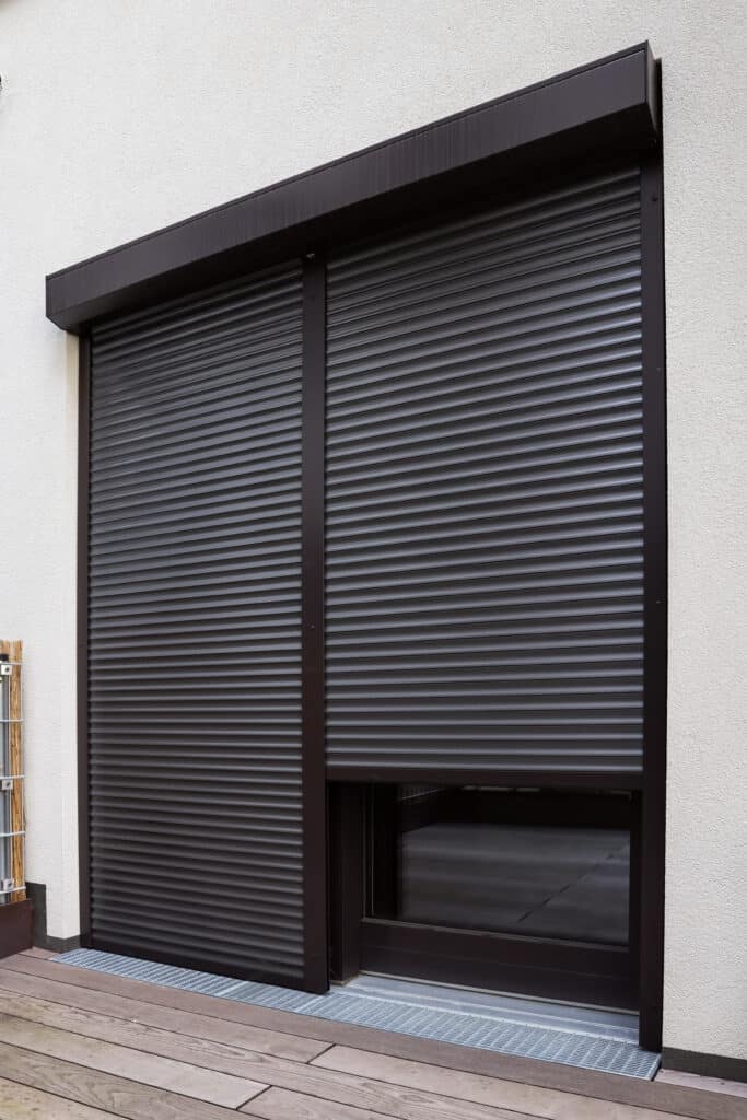 Façade moderne avec volet roulant marron recouvrant une porte-fenêtre. Terrasse en bois et grille de drainage.