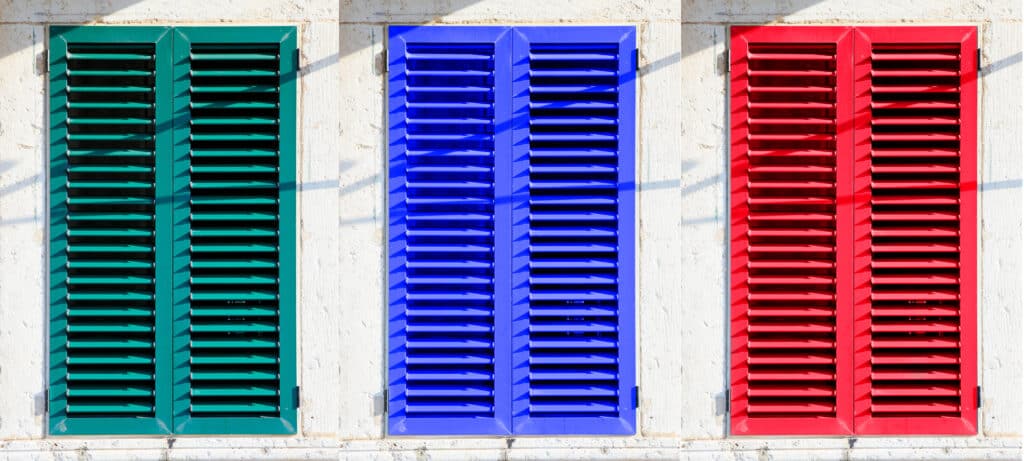 Trois volets persiennes extérieurs aux couleurs vives : vert sarcelle, bleu roi et rouge vif, devant un mur blanc texturé en plein soleil.